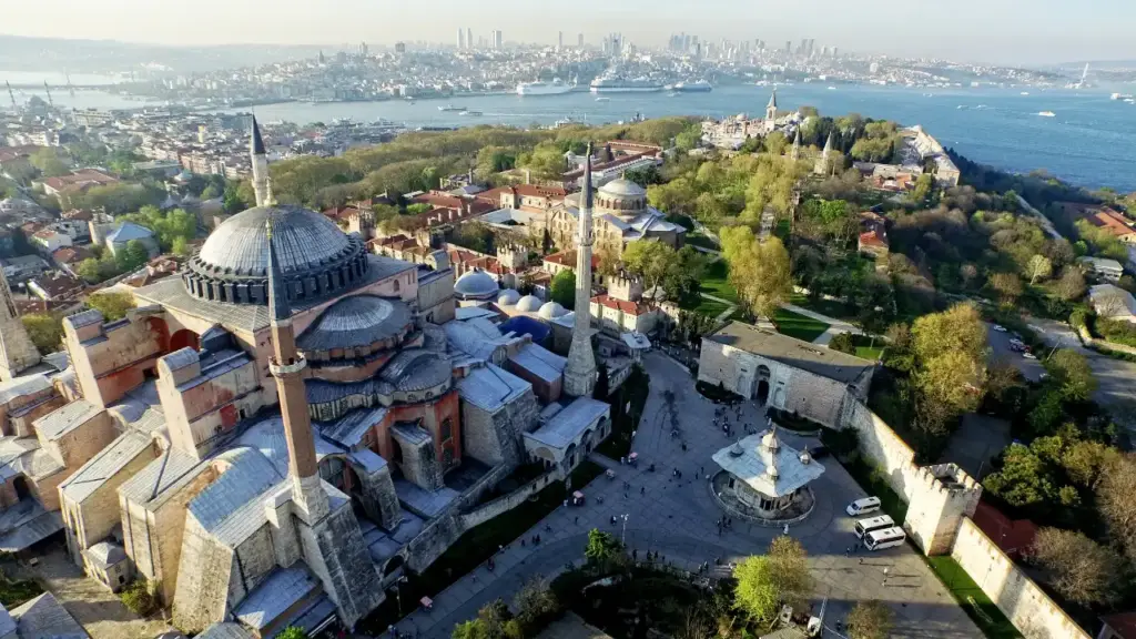 Aerial view of Hagia Sophia with its massive dome and four minarets, the entrance to Topkapı Palace, and the Bosphorus strait with the modern Istanbul skyline in the background