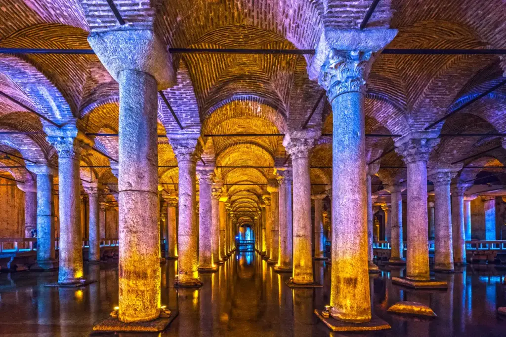 Inside the Basilica Cistern showing rows of marble columns reflected in shallow water, lit by blue and golden atmospheric lighting beneath brick vaulted ceilings