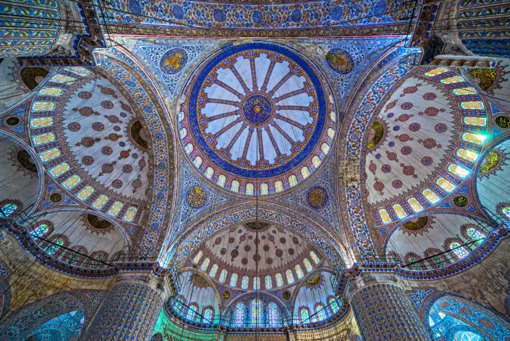 The painted ceiling of the Blue Mosque showing the central dome surrounded by four semi-domes, with blue and gold floral motifs, calligraphic medallions, and stained glass windows