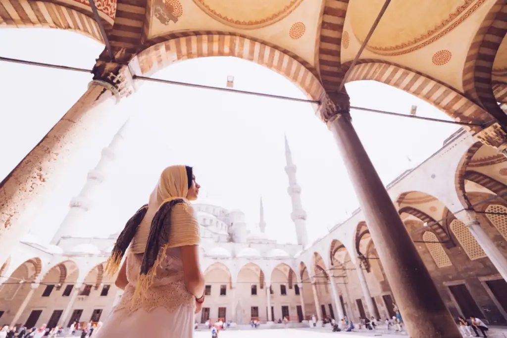 A visitor wearing a headscarf looking out across the Blue Mosque courtyard from beneath the domed colonnade, with the minarets and arched arcade visible beyond