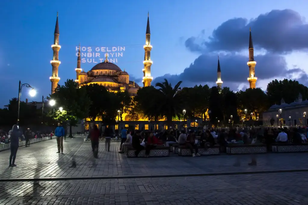 The Blue Mosque at dusk during Ramadan with a mahya light message strung between the minarets reading Hoş Geldin On Bir Ayın Sultanı, and crowds gathering in Sultanahmet Square