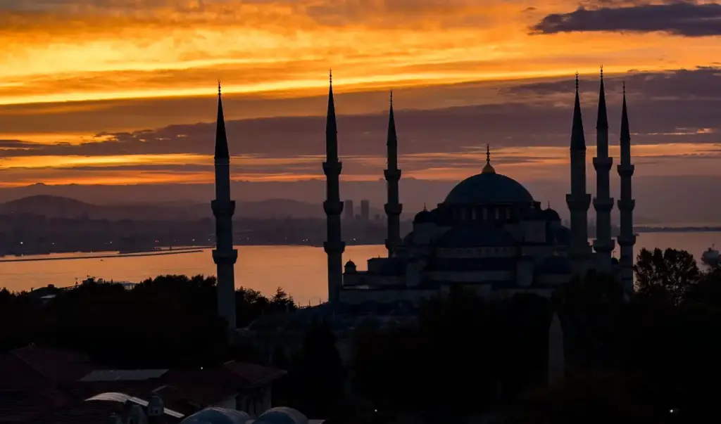 The Blue Mosque silhouetted against a dramatic orange and red sunset sky, with all six minarets visible and the Sea of Marmara and Asian shore in the background