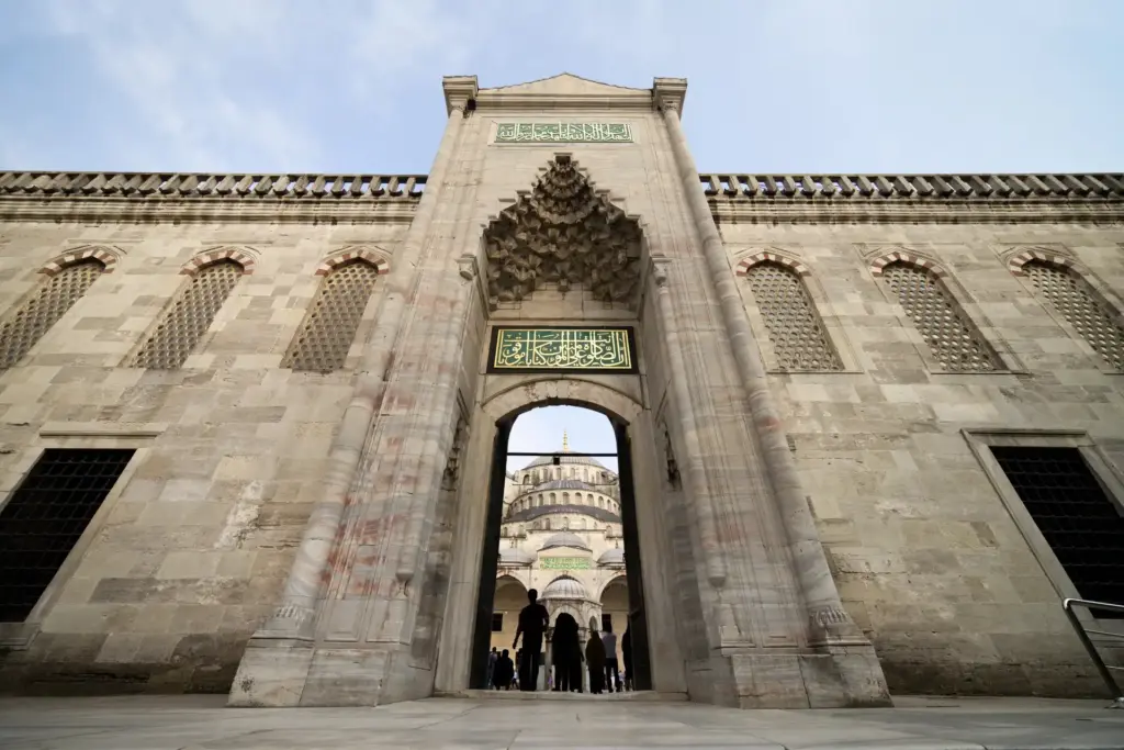 The main entrance gate of the Blue Mosque with its tall stone portal, muqarnas honeycomb vault, green calligraphic inscription panels, and visitors walking through into the courtyard with the mosque domes visible beyond