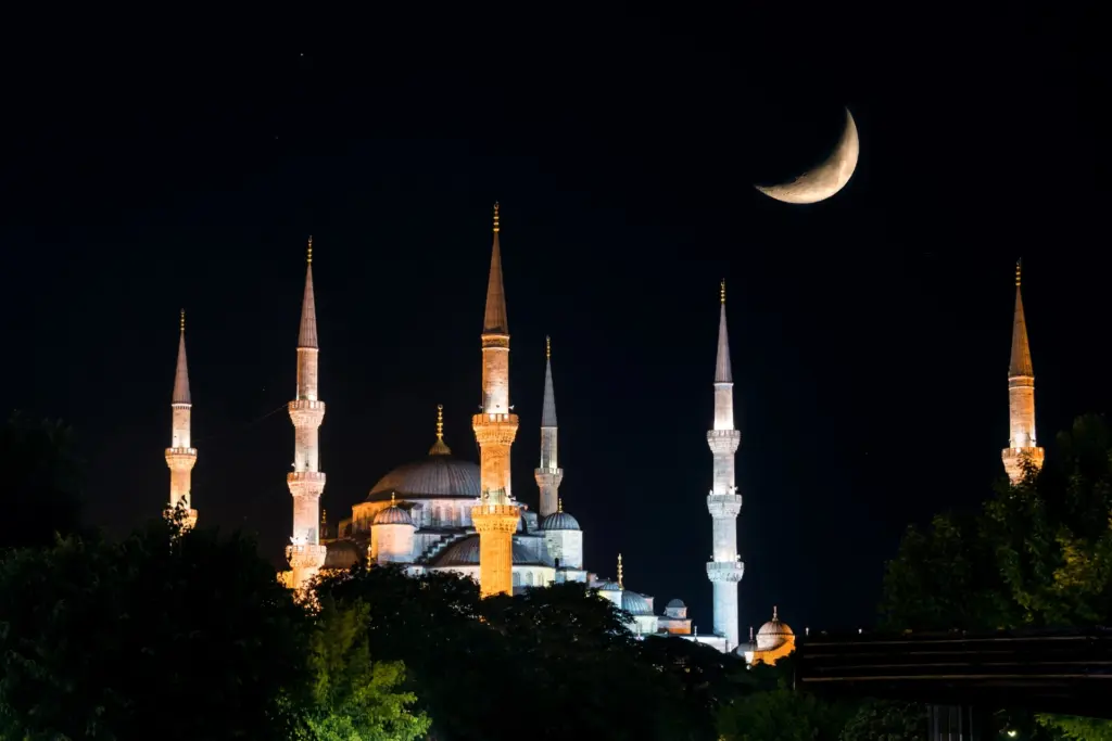 The Blue Mosque at night with all six illuminated minarets and a crescent moon in the dark sky above the domes