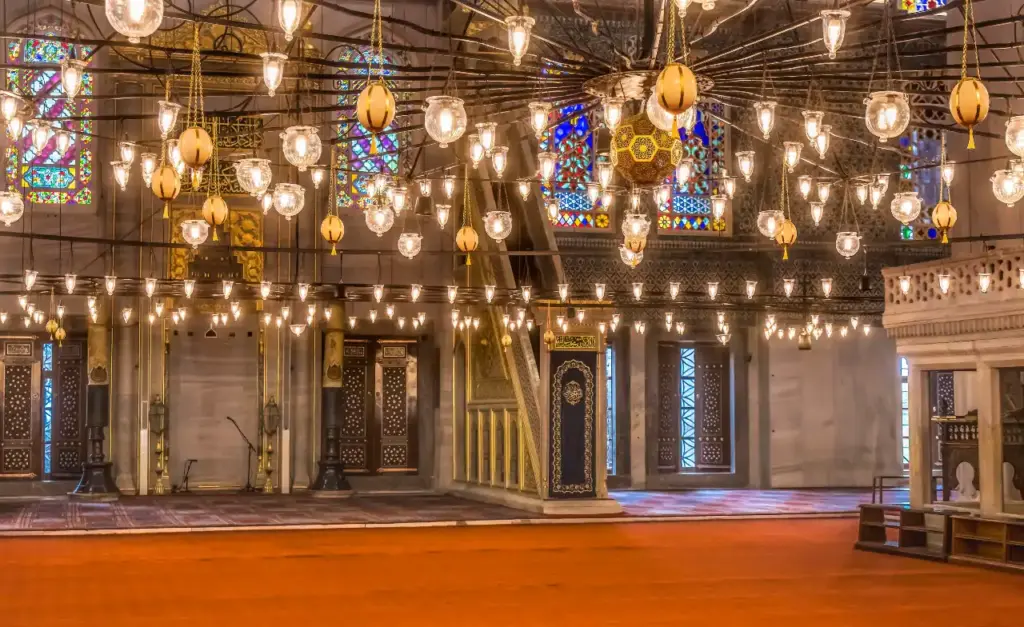 The mihrab and minbar of the Blue Mosque seen at floor level beneath rows of hanging glass lamps, with stained glass windows and the red carpet of the prayer hall in the foreground