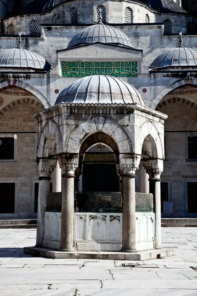 Close-up of the hexagonal ablution fountain in the Blue Mosque courtyard, with carved stone arches, marble columns, and a green calligraphic inscription panel on the mosque wall behind