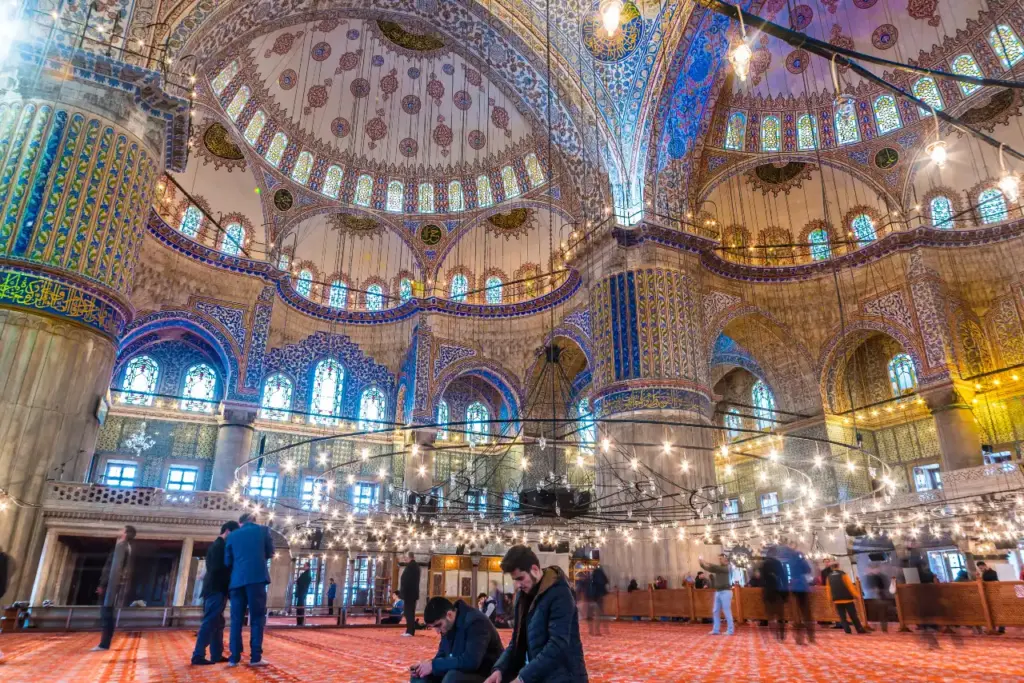 Visitors inside the Blue Mosque prayer hall with the blue İznik tile walls, cascading domes, circular chandelier, stained glass windows, and red carpet visible throughout the space
