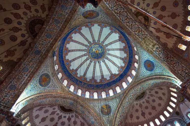 Looking up at the central dome of the Blue Mosque, showing the painted calligraphic medallions, radiating geometric patterns, and blue İznik tilework on the pendentives