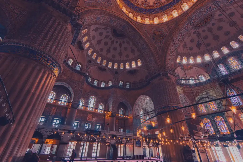 Inside the Blue Mosque prayer hall showing the cascading domes, stained glass windows, hanging chandeliers, and massive elephant foot pillars