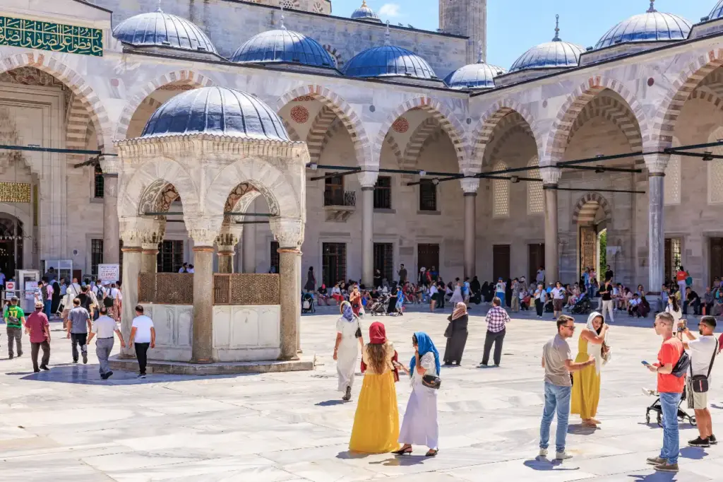 A busy summer day in the Blue Mosque courtyard with visitors and families walking past the ablution fountain, headscarves worn by women, and the domed colonnade and arched façade behind