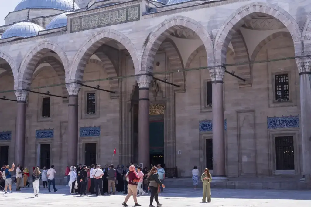 Visitors gathering at the courtyard entrance of the Blue Mosque, with the arched façade, blue İznik tile panels, green calligraphic inscription, and lead-covered domes above