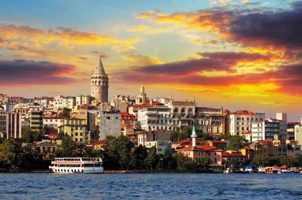 The Galata Tower and Beyoğlu district seen across the Golden Horn at sunset, with colourful buildings, ferry boats, and a dramatic orange sky