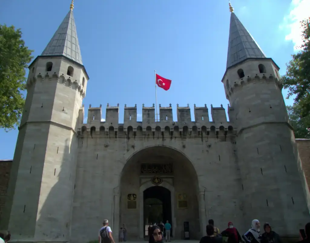 The Gate of Salutation entrance to Topkapı Palace with its two octagonal towers, crenellated walls, Turkish flag, and visitors passing through the archway